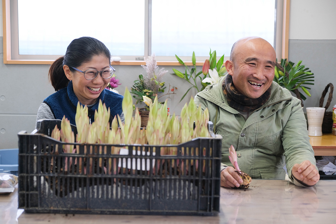 黒いプラスチックのカゴに入ったユリの苗の向こうに座る笑顔の野村英史さんとみゆきさんの画像