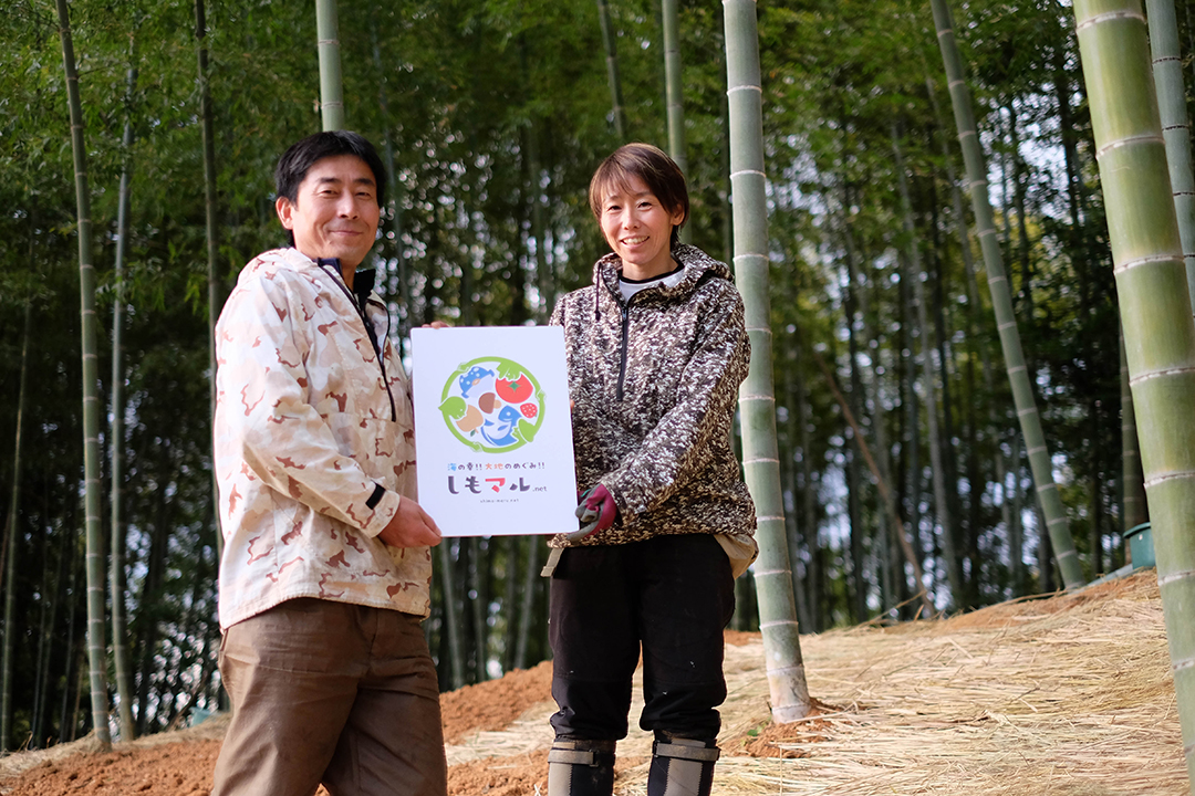 竹林でしもマルの看板を一緒に持って微笑んでいる山本由人さんと山本忍さんの画像