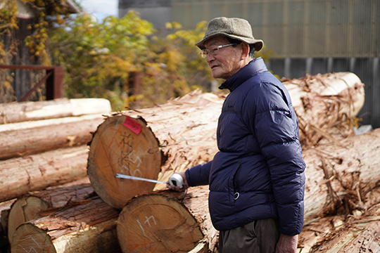 外の材木置き場で丸太の状態の木の断面をメジャーで指して話をしている松嵜行啓さんの画像