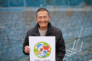 倉田博明さんが１人でナスの畑の前でしもマルの看板を持って笑っている画像