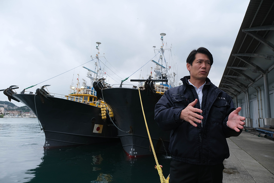 ２隻の沖合底引き網漁の船の前で両手を広げている宮本洋平さんの画像