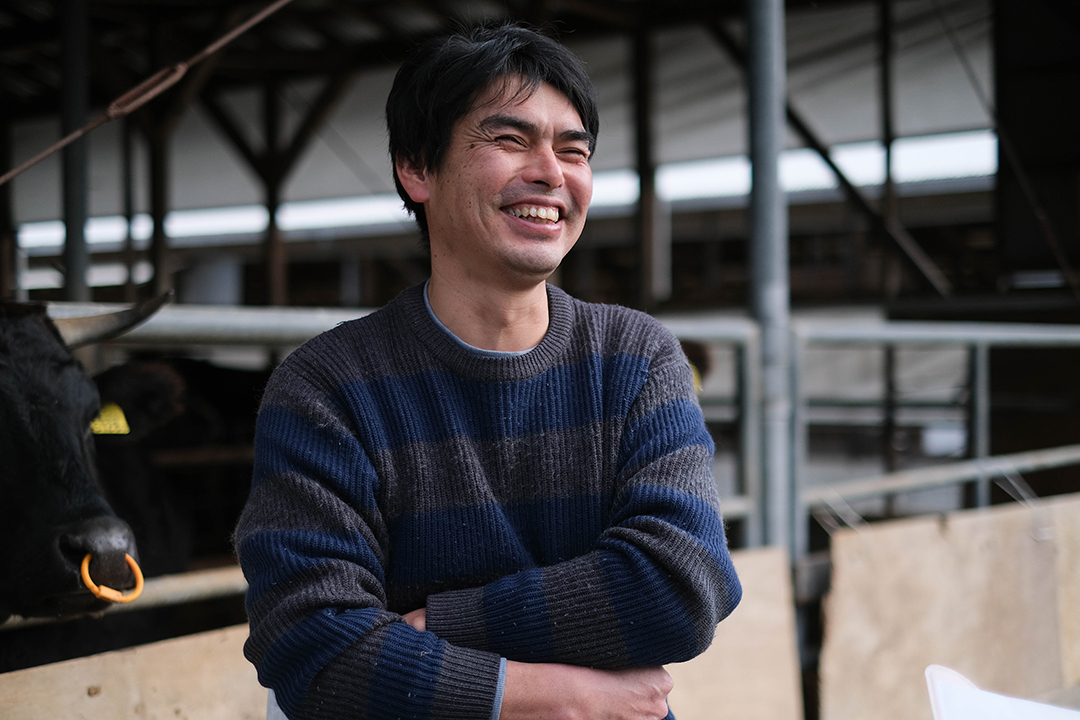 牛舎の前で腕を組んで笑っている吉冨貴博さんの画像