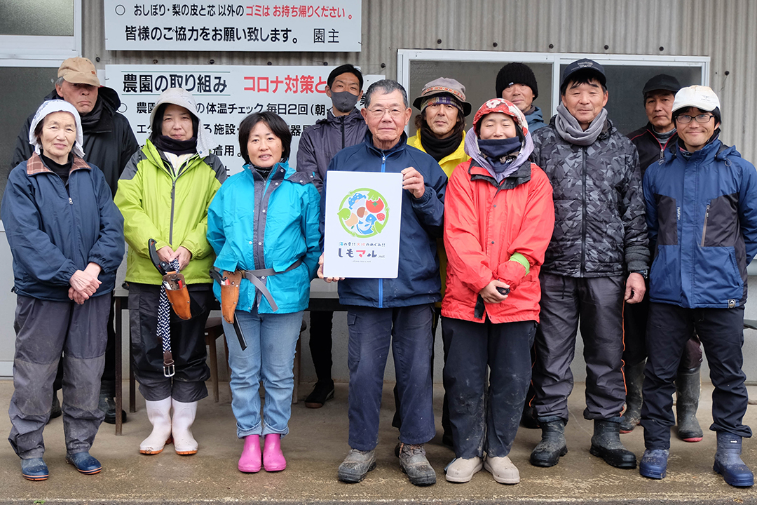 しもマルの看板を持つ河田征四郎さんと寺畑農園のスタッフの皆さんの画像