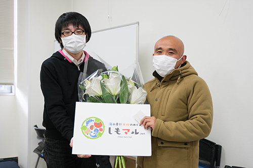 石川先生が生産者さんと花としもマルパネルを持って笑っている画像