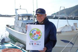 船の前でしもマルの看板を持って笑っている山口県漁業協同組合の伊東祥さんの画像