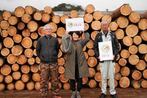 積み上げられた原木の切り口が並ぶ前に中野さんと山下さんと水上さんが並んで笑顔を見せている画像