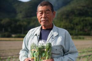 長岡明敏さんがはなっこりー畑の前ではなっこりーの袋を持って立っている画像